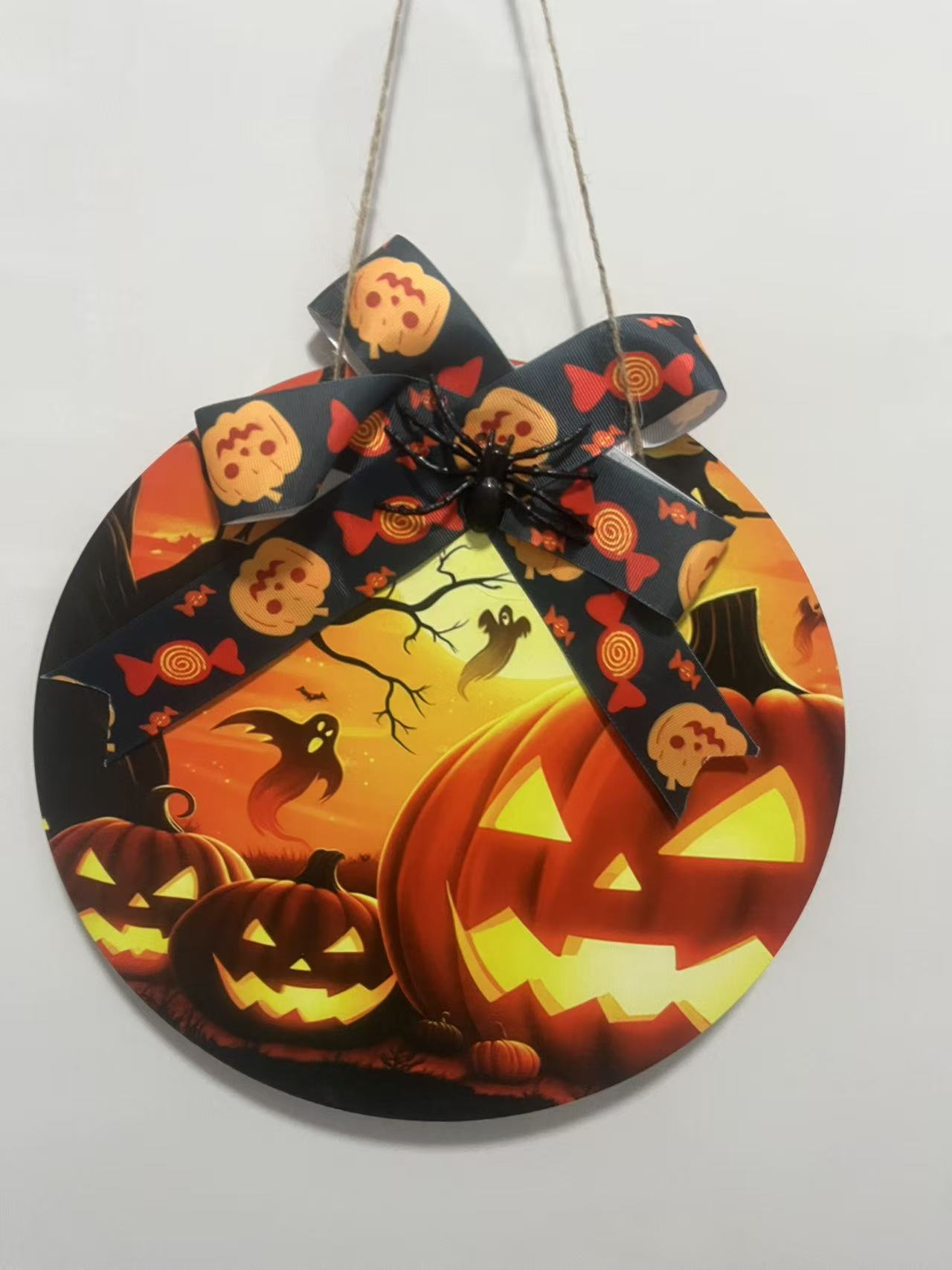 A circular Halloween hanging sign featuring vibrant Jack-O-Lantern and ghost illustrations, topped with a decorative pumpkin-patterned bow and a black spider ornament, suitable for front door holiday decor.